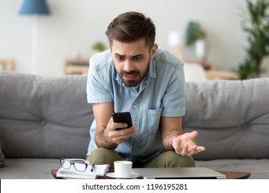 Confused millennial man sitting alone on couch in living room at home looking at smartphone screen with irritation and indignance. Annoyed male received bad news, device gadget broken or dead concept - Shutterstock ID 1196195281