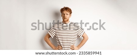 Confused and offended man with ginger curly hair staring at camera puzzled, standing questioned against white background.