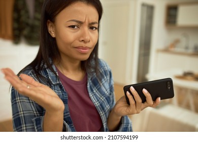 Confused Pretty Dark-skinned Woman Holding Smartphone, Shrugging After Conversation With Stranger, Looking At Camera Frowning, Tighten Lips. Human Emotions, Feelings, Gestures, Signs. Body Language