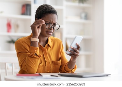 Confused pretty millennial black woman in casual outfit looking at cell phone screen and touching her eyeglasses, checking exciting online offer, reading weird text, copy space, office interior