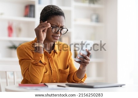 Confused pretty millennial black woman in casual outfit looking at cell phone screen and touching her eyeglasses, checking exciting online offer, reading weird text, copy space, office interior