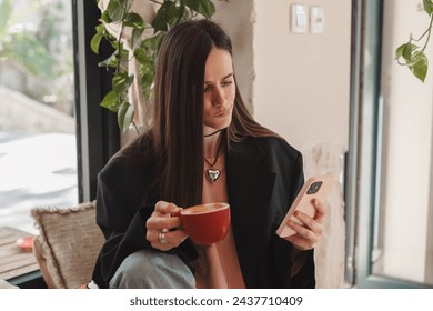 Confused puzzled brunette woman in casual clothes, sits in cafe, holds a smartphone in her hand, looks questioningly, unhappy. Girl getting surprising bad news, annoyed woman think about solutions.