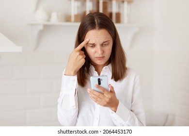 Confused Puzzled Dark Haired Woman Standing Cell Phone In Hands, Checking Social Networks, Reading Negative Message Or Comment, Posing In The Kitchen At Home.