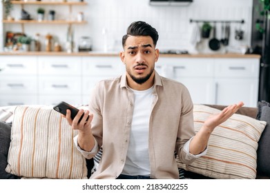 Confused puzzled indian or arabian guy in casual clothes, sits on a sofa in an interior living room, holds a smartphone in his hand, looks questioningly at the camera, spreading his arms around