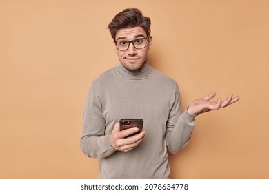 Confused questioned young man shrugs shoulders wears optical glasses and turtleneck holds mobile phone reads something strange browses newsfeed via social networks puzzled about online content
