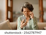 Confused senior woman in eyeglasses squinting eyes, try to read message on cellphone, having problems with vision, sitting on couch at home