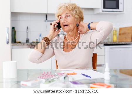 Confused senior woman sitting at kitchen table among a lot of medicines and talking on phone