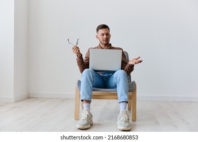 Confused Shocked Irritated Young Tanned Man With Laptop On Knees Can Not Understand Work Task Computer Error Say What Sinning On Chair At White Home Wall. Shocked By Gadget Breakdown Offer. Copy Space