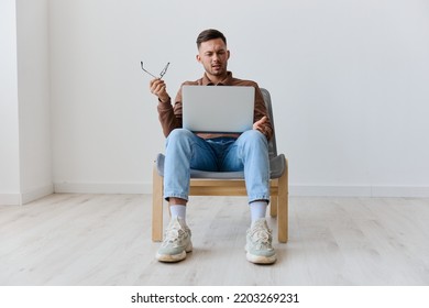 Confused Shocked Irritated Young Tanned Man With Laptop On Knees Can Not Understand Work Task Computer Error Say What Sinning On Chair At White Home Wall. Shocked By Gadget Breakdown Offer. Copy Space