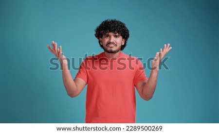 confused or shocked young adult man with beard wearing coral t-shirt standing with raised arms and looking at camera asking. Isolated on a blue studio background.