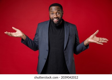 Confused Smiling Smirked Puzzled Young African American Business Man 20s Wearing Classic Jacket Suit Standing Spreading Hands Looking Camera Isolated On Bright Red Color Background Studio Portrait