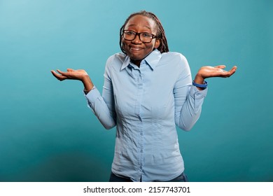 Confused And Uncertain Woman Shrugging Shoulders With Uncertainty While Standing On Blue Background. Puzzled And Doubtful Young Adult Person Being Hesitant And Unconfident While Answering Question.