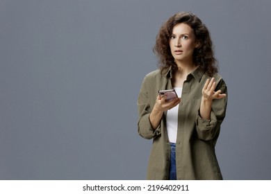 Confused Unhappy Angry Curly Beautiful Woman In Casual Khaki Green Shirt Get Fake Message From Frauds Using Phone Posing Isolated On Gray Blue Background. Social Media Influencer Concept. Copy Space