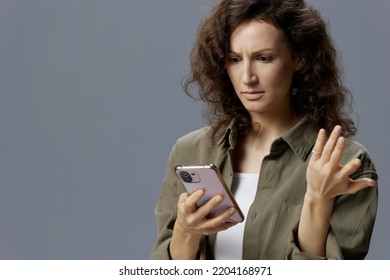 Confused Unhappy Angry Curly Beautiful Woman In Casual Khaki Green Shirt Get Fake Message From Frauds Using Phone Posing Isolated On Gray Blue Background. Social Media Influencer Concept. Copy Space