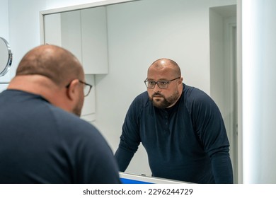 Confused upset overweight middle aged man looking at mirror in bathroom at home. Bearded male in eyeglasses dissatisfied with appearance. Midlife crisis, psychological problems, depression concept. - Powered by Shutterstock