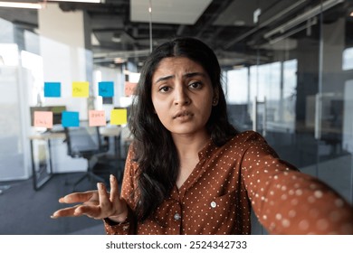 Confused woman in office making video call using phone. Businesswoman surrounded by colorful sticky notes on glass wall during brainstorming. Young professional in casual office expressing concern. - Powered by Shutterstock