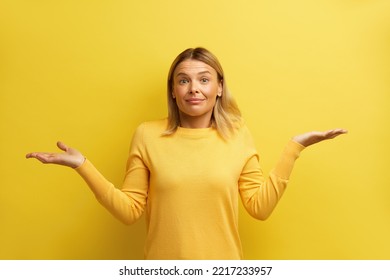 Confused Woman Shrugging Hands. Portrait Of Uncertain Girl Expressing Doubts And Bewilderment, Looking At Camera With Question So What, Who Cares. Indoor Studio Shot Yellow Background 