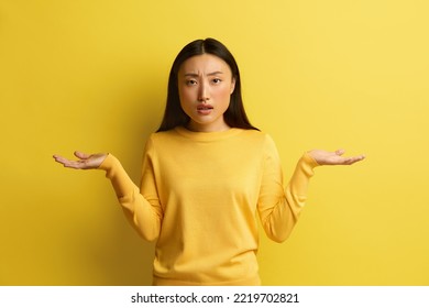 Confused Woman Shrugging Hands. Portrait Of Uncertain Asian Girl Expressing Doubts And Bewilderment, Looking At Camera With Question So What, Who Cares. Indoor Studio Shot Yellow Background 