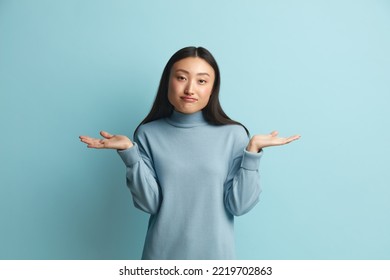 Confused Woman Shrugging Hands. Portrait Of Uncertain Asian Girl Expressing Doubts And Bewilderment, Looking At Camera With Question So What, Who Cares. Indoor Studio Shot Blue Background 