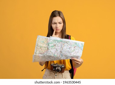Confused woman traveler with camera looking at city map, got lost, yellow studio background, copy space. Upset young lady backpacker lost in city, checking map. Navigation, trekking during travelling - Shutterstock ID 1963530445