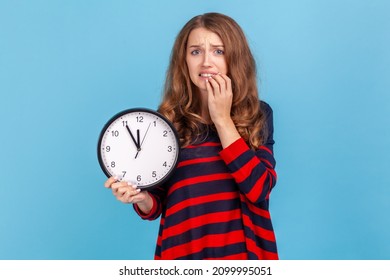 Confused woman wearing striped casual style sweater, holding wall clock in hands, biting nails, looking at camera with nervous expression, deadline. Indoor studio shot isolated on blue background.