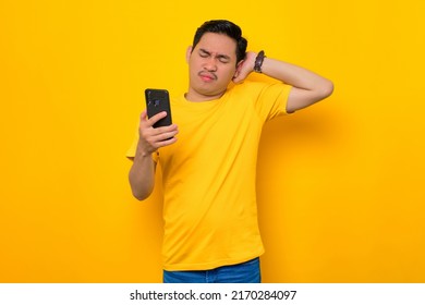 Confused Young Asian Man In Casual T-shirt Using Mobile Phone While Scratching Head, Reacting To Online News Isolated On Yellow Background. People Lifestyle Concept