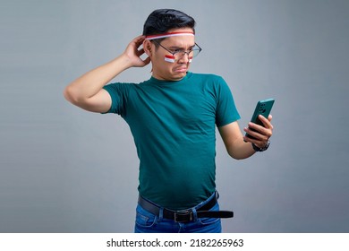 Confused Young Asian Man In Casual T-shirt  Scratching Head While Using Mobile Phone, Worried About Something Isolated On Grey Background. Indonesian Independence Day Celebration Concept