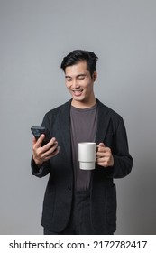 Confused Young Asian Man Holding Mobile Phone And Feeling Sleepy, Lonely On Gray Background.