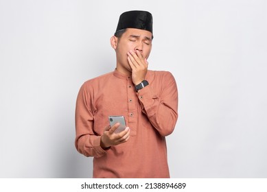 Confused young Asian Muslim man holding mobile phone and feeling sleepy isolated over white background