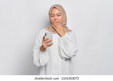 Confused young Asian Muslim woman holding mobile phone and feeling sleepy isolated over white background