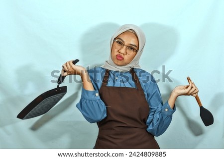 confused young Asian Muslim woman housewife wearing brown apron and glasses holding spatula and pan while doing housework isolated onwhite background. muslim housewife concept