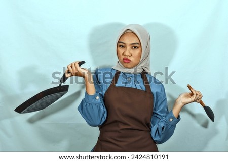 confused young Asian Muslim woman housewife wearing brown apron holding spatula and pan while doing housework isolated onwhite background. muslim housewife concept
