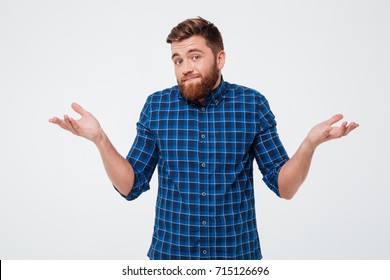 Confused young bearded man standing and shrugging shoulders isolated over white background