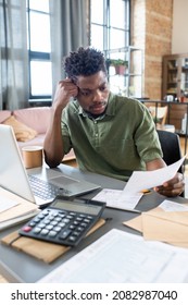 Confused Young Blackman In Green Shirt Sitting At Desk With Laptop And Examining Financial Papers