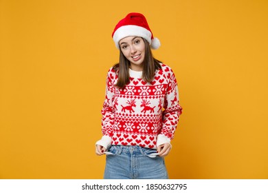 Confused Young Brunette Santa Woman In Red Sweater, Christmas Hat Has No Money Showing Empty Pockets Isolated On Yellow Background, Studio Portrait. Happy New Year Celebration Merry Holiday Concept