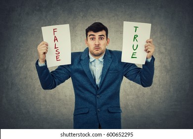 Confused Young Businessman Holding Two White Papers Try To Choose Between True Or False, Grey Wall Background. Human Emotion, Face Expression, Life Perception Concept.