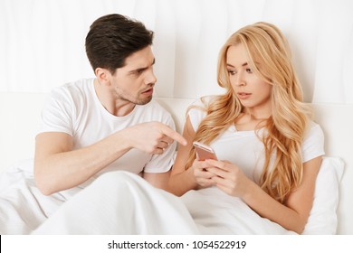 Confused Young Couple Having An Argument About Mobile Phone While Lying In Bed At Home