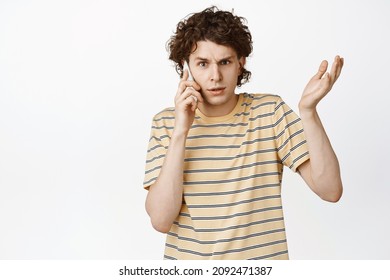 Confused Young Man Having A Strange Phone Call, Shrugging And Looking Puzzled, Talking On Mobile, Standing Over White Background