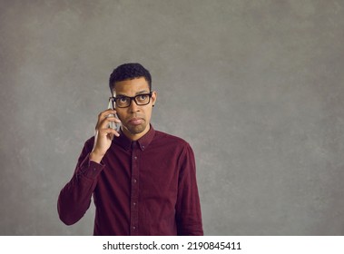 Confused Young Man Making A Call To Request Help At Customer Support Center. Annoyed Discontent Black Guy Holding Mobile Phone To His Ear Waiting For Answer Standing Against Grey Copyspace