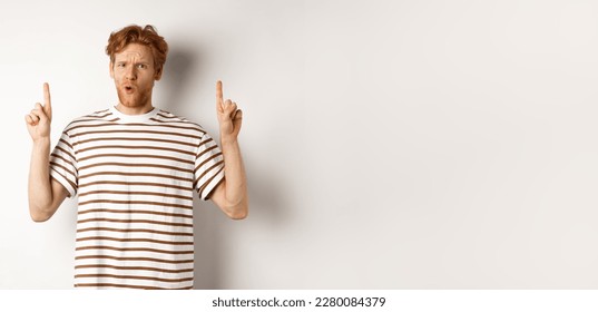 Confused young man with red curly hair pointing fingers up, staring shocked and puzzled, standing over white background. - Powered by Shutterstock