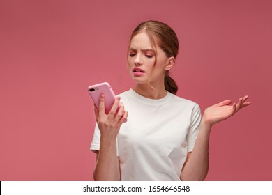 Confused young redhead woman holding pink smartphone.Embarassed girl has problem with broken not working mobile phone.Receiving bad news or email,spam message.Young people working with mobile devices.