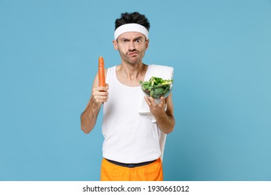 Confused Young Sporty Fitness Man With Thin Skinny Body Sportsman In Headband Shirt Shorts Hold Vegetable Salad In Glass Bowl Carrot Isolated On Blue Background. Workout Gym Sport Motivation Concept