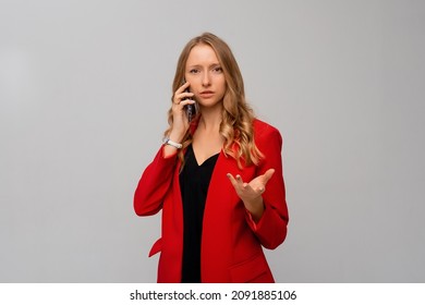 Confused Young Woman Having A Strange Phone Call, Shrugging And Looking Puzzled, Talking On Mobile, Standing In Red Blazer Against Gray Background