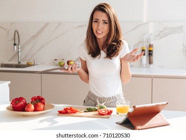 Confused young woman holding knife and shrugging shoulders while cooking on a kitchen - Shutterstock ID 710120530