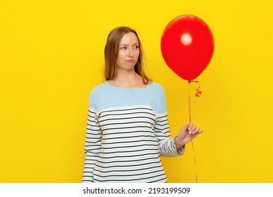 Confused Young Woman Holds Red Inflated Helium Balloon, Feels Embarrassed And Offended Has Bad Mood During Celebration. Holiday And Party Concept