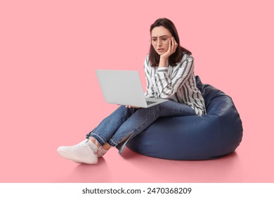 Confused young woman with laptop sitting on bean bag chair against pink background - Powered by Shutterstock