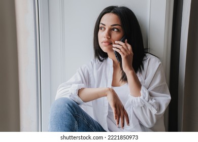 Confused Young Woman In White Shirt And Blue Jeans Sitting On Windowsill Looking Outside Feels Loneliness Talking By Phone. Tired African American Girl Using Cellphone, In Troubles, Divorce, Mistake.