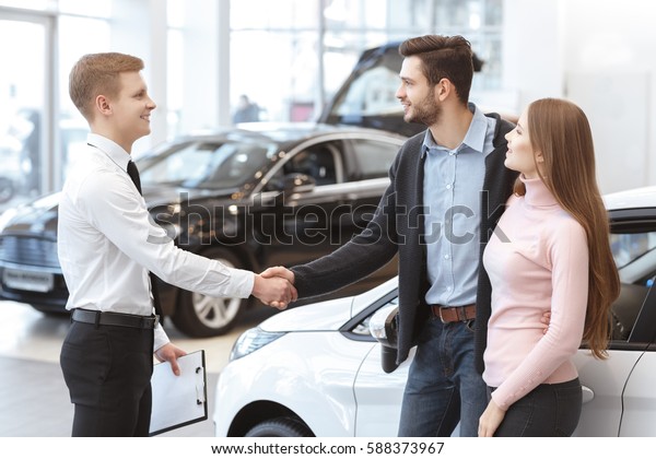Congrats with your new car! Happy young couple\
buying a new car at the dealership handsome man shaking hands with\
a car dealer hugging his beautiful girlfriend family lifestyle\
luxury rental concept