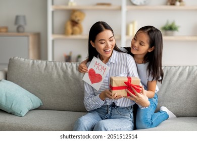 Congratulations. Loving Asian Daughter Greeting Surprised Mom With Mother's Day, Giving Her Handmade Card And Gift Box, Sitting On Sofa At Home Interior, Free Space