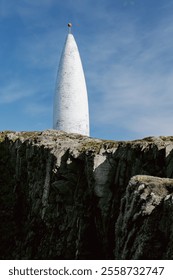 A conical, white navigation marker is positioned on a rugged, dark clifftop. The day is sunny and the sky is mostly clear.